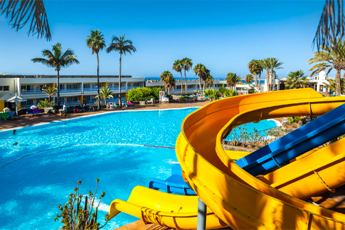 Gele en blauwe waterglijbaan die uitkomt in een groot zwembad, met palmbomen en het hotel op de achtergrond bij Abora Interclub Atlantic.